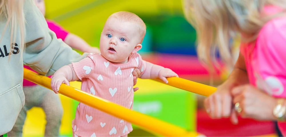 An infant wearing a pink outfit with heart patterns holds onto a yellow bar for balance, supported by two adults in a vibrant play area.