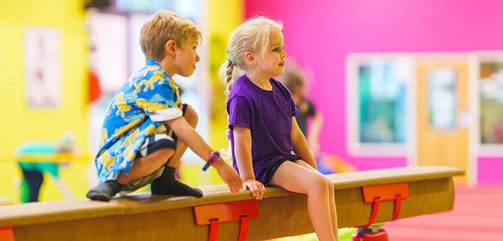 Two young children sit on a balance beam, one wearing a purple shirt and the other wearing a blue shirt with a yellow animal pattern, in a brightly colored gym.