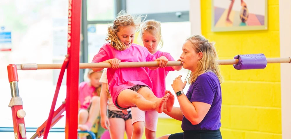 A young girl wearing a pink shirt is assisted on the uneven bars by a coach in a purple shirt, with other children waiting their turn in the background.