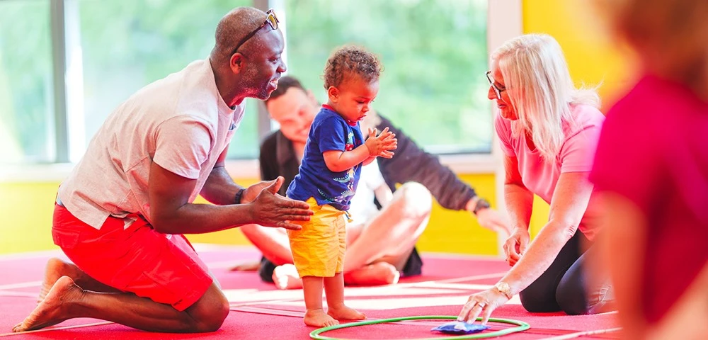 A young child claps while standing on a red mat, guided by an adult male and Yvonne in a gym-like setting with bright yellow walls.