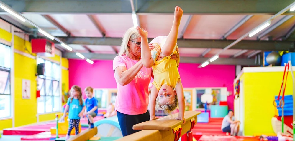 A child performs a handstand on a balance beam, assisted by Yvonne wearing a pink shirt, surrounded by other children in a colourful gym